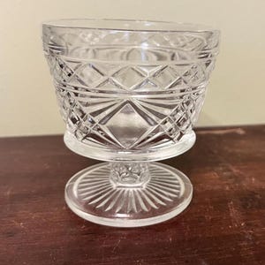 May include: A clear, vintage-style glass dessert dish with a diamond pattern on the bowl and a circular base. The stem is short and the base is decorated with radiating lines. The dish is likely made of pressed glass.
