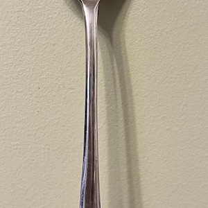 May include: A silver serving spoon with a distinctive three-pronged head. The spoon's long handle tapers to a point and features an engraved initial. The spoon is likely used for serving salads or other dishes.