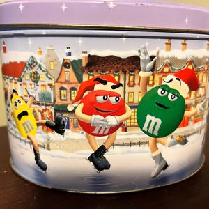 Może przedstawiać: Okrągłe, metalowe pudełko z fioletową pokrywką i zimową sceną. Pudełko przedstawia trzy postacie M&M's w czapkach Świętego Mikołaja, które jeżdżą na łyżwach po ośnieżonej ulicy. Postacie są czerwone, zielone i żółte.