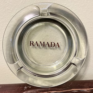 May include: Circular clear glass ashtray with three cigarette rests. The ashtray has the word "RAMADA" printed in dark red in the centre. The ashtray is designed for use with cigarettes.