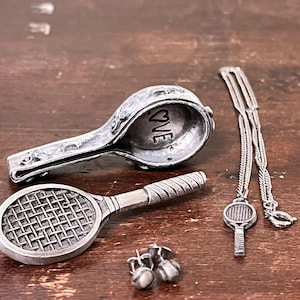 May include: A collection of silver tennis-themed jewelry and accessories. Includes a tennis racket pendant necklace, tennis ball earrings, and a tennis racket-shaped container with the word "LOVE" inside. The items are displayed on a wooden surface.
