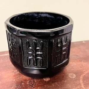 May include: A black, octagonal-shaped decorative container. The container has a glossy finish and features embossed geometric patterns around the exterior. The container sits on a wooden surface.