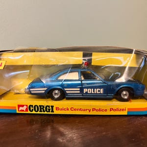 Puede incluir: Un coche de policía Corgi Buick Century azul en su embalaje original. El coche de juguete tiene la palabra "POLICE" impresa en el lateral. El embalaje es amarillo, azul y naranja con la palabra "NEW" en la parte superior izquierda.