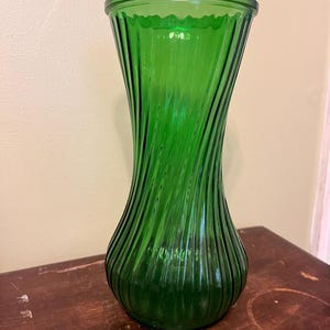 Puede incluir: Un jarrón de vidrio verde con un diseño acanalado en espiral. El jarrón tiene una parte superior ancha y acampanada y una base redondeada. El vidrio es de un tono verde vibrante, y el jarrón está sobre una superficie de madera oscura.