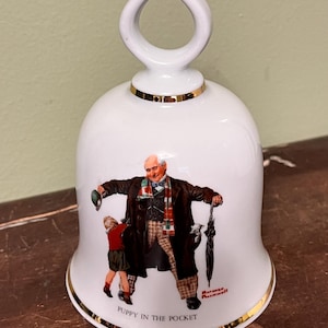 May include: White ceramic bell with a gold rim and handle. The bell features a colorful illustration of a man and child with the text "PUPPY IN THE POCKET" and the artist's signature. The bell is approximately 6 inches tall.