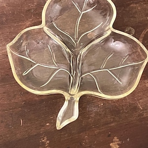 Puede incluir: Un plato de servir transparente en forma de hoja con tres secciones. El plato tiene un borde amarillo claro y patrones detallados de venas de hojas. El plato está diseñado para contener aperitivos o artículos pequeños. El plato está sobre una superficie de madera marrón oscuro.