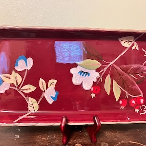 May include: Rectangular serving tray with a deep red background and floral design. The tray features white and blue flowers, green leaves, and red berries. The rim has a textured, light brown border.