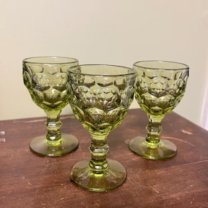 May include: Three green-tinted, vintage-style goblets. Each glass features a textured, honeycomb-like pattern on the bowl and a detailed stem. The glasses are displayed on a dark wooden surface, showcasing their unique design.