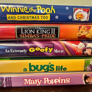 May include: A stack of VHS tapes featuring Disney animated movies. Titles include "Winnie the Pooh and Christmas Too," "The Lion King II: Simba's Pride," "An Extremely Goofy Movie," "a bug's life," and "Mary Poppins."