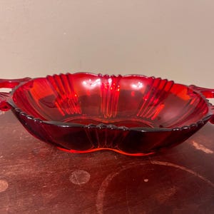 May include: A translucent ruby red glass dish featuring an oval shape with scalloped edges and two integrated handles on opposite sides. The interior surface displays a radiating ribbed pattern that extends from the center toward the rim.
