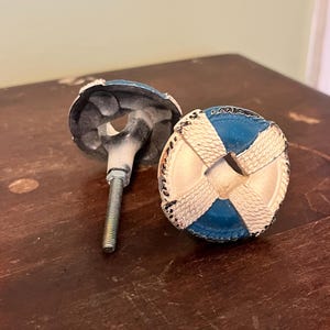 以下が含まれることがあります： 2つの装飾的なキャビネットノブ。各ノブは、青い中心と白い十字模様の円形デザインで、救命浮輪に似ています。ノブは金属仕上げで、取り付け用のねじ付きボルトが付いています。