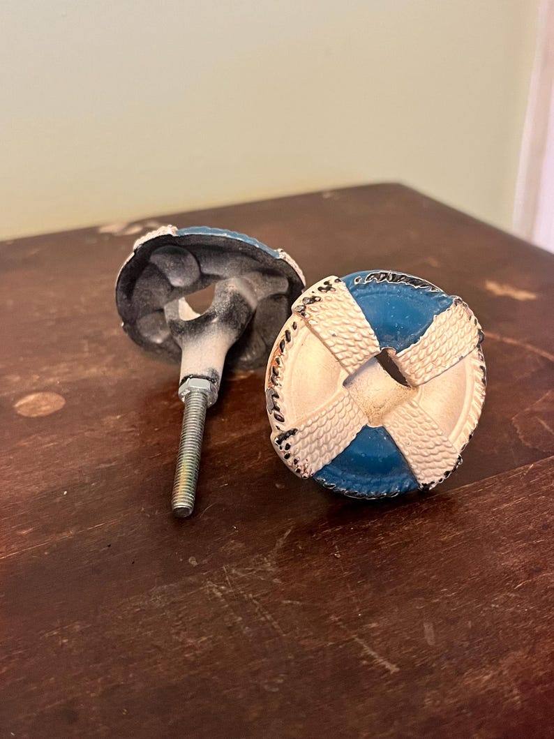 以下が含まれることがあります： 2つの装飾的なキャビネットノブ。各ノブは、青い中心と白い十字模様の円形デザインで、救命浮輪に似ています。ノブは金属仕上げで、取り付け用のねじ付きボルトが付いています。