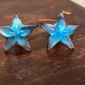 以下が含まれることがあります： 2つの装飾ガラス星オーナメント。各星は、テクスチャ加工された黒い先端を持つ鮮やかな青色の中心を持っています。星は濃い茶色の木製の表面に表示されています。