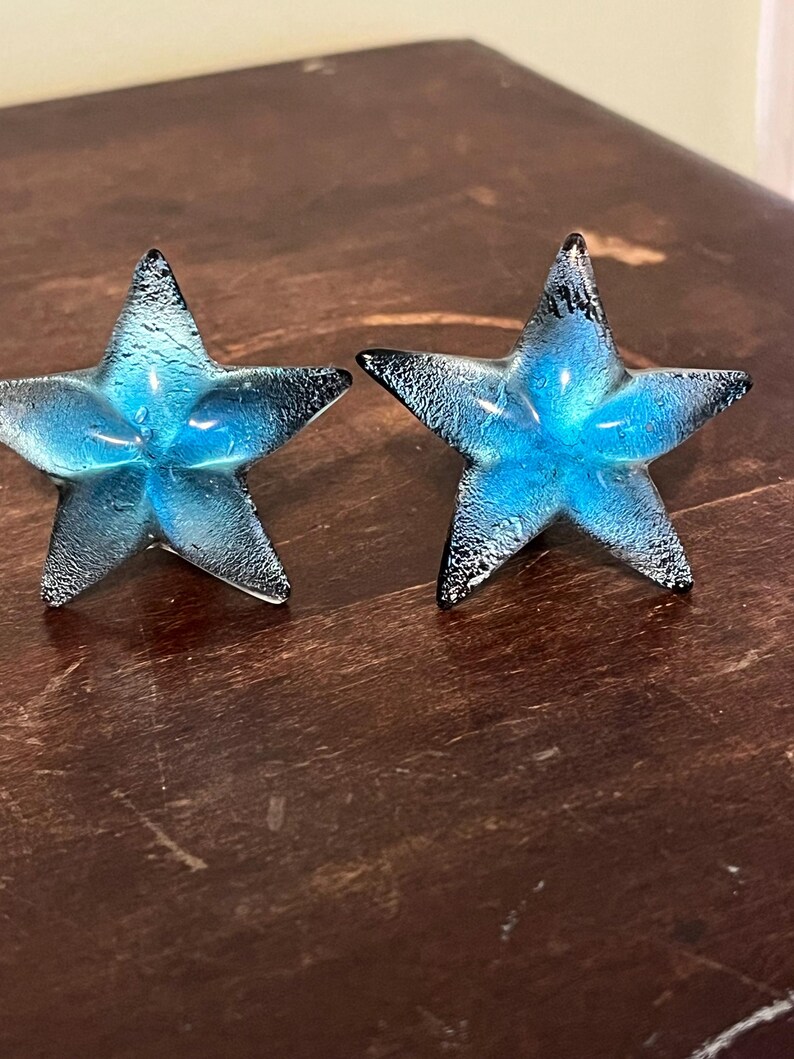 以下が含まれることがあります： 2つの装飾ガラス星オーナメント。各星は、テクスチャ加工された黒い先端を持つ鮮やかな青色の中心を持っています。星は濃い茶色の木製の表面に表示されています。