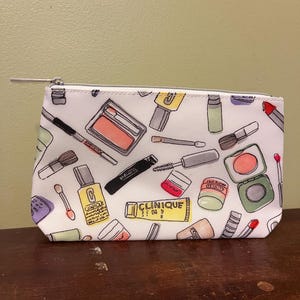 May include: White cosmetic bag with a silver zipper, patterned with colorful makeup items. The bag features illustrations of makeup brushes, eyeshadow, mascara, lipstick, and bottles of Clinique products.