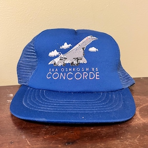 May include: A blue trucker hat with a white and gray graphic of a Concorde airplane and the text "EAA OSHKOSH '85 CONCORDE". The hat has a mesh back and a curved brim.