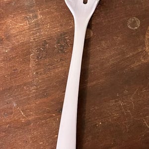 May include: A white spaghetti server with a slotted head and long handle. The head has multiple tines for grabbing pasta, and a central slot for draining. The handle has a hole for hanging. Kitchen utensil.