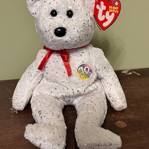 Könnte beinhalten: Ein weißer Beanie Baby-Bär mit silbernen Glitzerakzenten. Der Bär hat schwarze Augen und eine schwarze Nase sowie ein rotes Band. Ein Etikett zeigt "10 Year Anniversary" und "Beanie Babies". Ein Sammler-Stofftier.