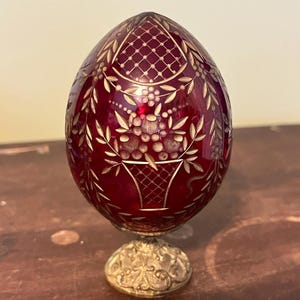 Puede incluir: Un objeto decorativo en forma de huevo, de color rojo rubí, con diseños florales y de celosía grabados en oro. El huevo se apoya sobre un soporte de metal dorado y detallado. La superficie del huevo refleja la luz, resaltando los intrincados patrones.