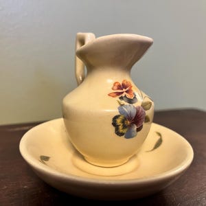 May include: A small, cream-coloured ceramic jug and bowl set. The jug has a handle and is decorated with floral designs in shades of orange, purple, and blue. The bowl has a matching floral design.