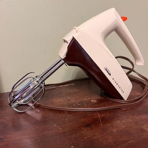May include: Vintage Sunbeam Mixmaster hand mixer in cream and brown. The mixer has a brown handle and a cream-colored body with the brand name printed on the side. The beaters are made of stainless steel. The power cord is brown.