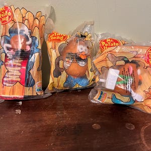 Puede incluir: Varias figuras de juguete de Mr. Potato Head selladas en su embalaje original. El embalaje presenta el personaje de Mr. Potato Head con varios accesorios y el texto "Mr. Potato Head". Las bolsas son naranjas y amarillas.
