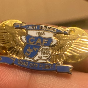 May include: Gold and blue pin with a winged design. The pin features the text "Ghost Squadron 1963", "CAF", and "Quad City Flight".