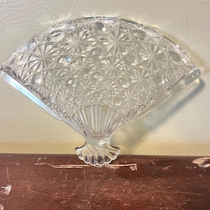 Può includere: Ventaglio decorativo in vetro trasparente con un motivo geometrico dettagliato. Il ventaglio ha un bordo smerlato e una base decorativa. Poggia su una superficie di legno marrone.