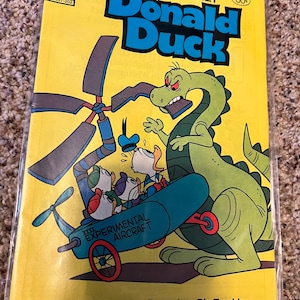 Könnte beinhalten: Ein Vintage-Comic-Cover mit Donald Duck und seinen Neffen in einem blauen und gelben Experimentalflugzeug, das von einem grünen Dinosaurier verfolgt wird. Der Text auf dem Cover lautet "Walt Disney Donald Duck" und "Donald und die Jungs geraten in große Schwierigkeiten auf der Suche nach dem GOLDENEN DINOSAURIER!"