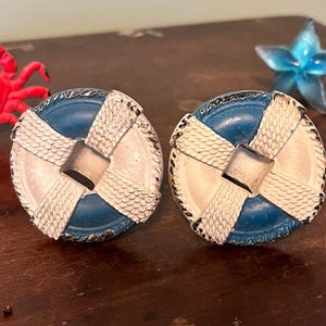 以下が含まれることがあります： 航海をテーマにしたカフスボタンのペア。各カフスボタンは、救命浮輪に似た青と白のデザインで、中央に銀色の正方形があります。カフスボタンは、ロープのような質感の縁取りが施されています。