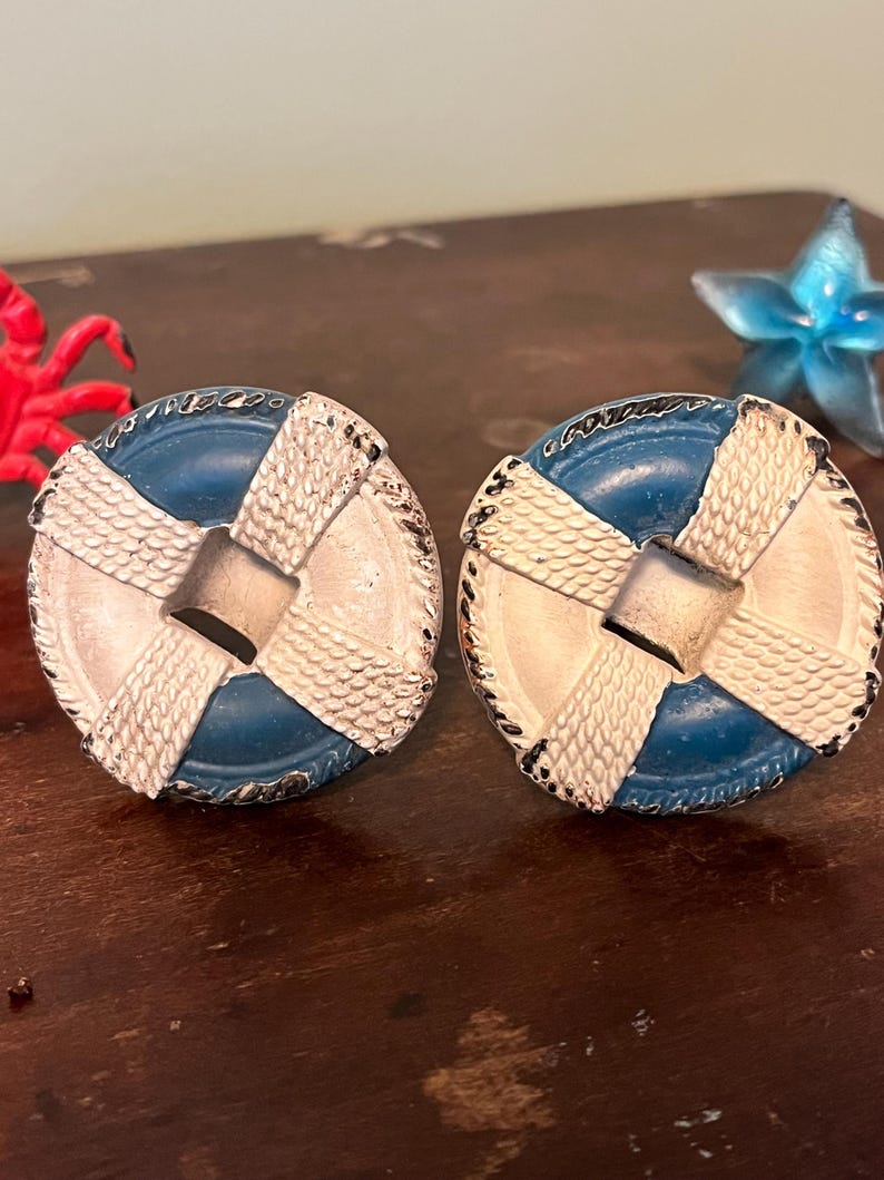 以下が含まれることがあります： 航海をテーマにしたカフスボタンのペア。各カフスボタンは、救命浮輪に似た青と白のデザインで、中央に銀色の正方形があります。カフスボタンは、ロープのような質感の縁取りが施されています。