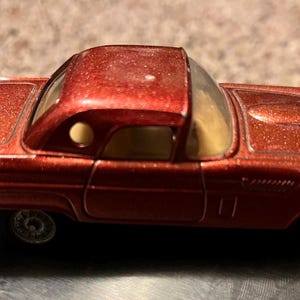 Puede incluir: Un coche de juguete rojo vintage con techo rígido. El coche tiene una rueda de repuesto en la parte trasera y ruedas plateadas. La carrocería del coche es de color rojo metalizado. Un modelo clásico.