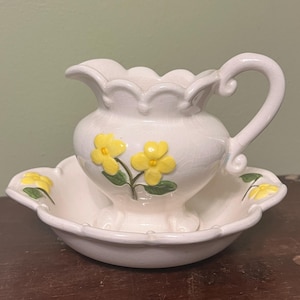 May include: A white ceramic jug and basin set. The jug has a curved handle and is decorated with yellow flower appliques. The basin has a scalloped edge and matching floral accents. The set is likely vintage.