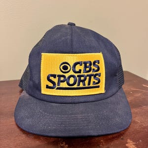 Könnte beinhalten: Marineblaue Trucker-Mütze mit einem gelben rechteckigen Aufnäher mit dem Text "CBS SPORTS" in Marineblau. Die Mütze hat einen gebogenen Schirm und eine Netzrückseite.