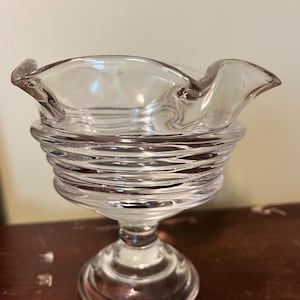 May include: A clear glass pedestal bowl with a ruffled rim. The bowl has a ribbed, cylindrical base and a rounded foot. The glass is transparent, allowing light to pass through. The bowl is likely vintage or antique.