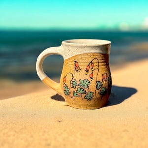 Peut inclure: Une tasse en céramique faite à la main avec une base beige et un bord et une anse blancs. La tasse est décorée d'un motif floral vert, orange et rouge. La tasse est posée sur une surface sablonneuse avec l'océan en arrière-plan.