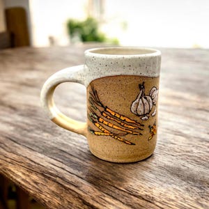 Peut inclure: Mug en céramique faite à la main, base beige et bord blanc moucheté. Le mug présente un motif peint à la main de carottes, d'ail et d'un piment. La anse est de couleur beige clair.