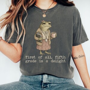 May include: A charcoal gray short-sleeve t-shirt featuring a graphic of a frog dressed in a beige cardigan and pink skirt, holding a notebook and coffee mug. The text below reads, first of all, fifth grade is a delight, in a typewriter font.
