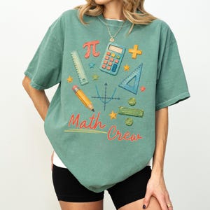 May include: A sage green short-sleeve t-shirt featuring a graphic print of school supplies including a calculator, ruler, pencil, and math symbols. The text "Math Crew" is printed in red and yellow below a blue coordinate plane graph.