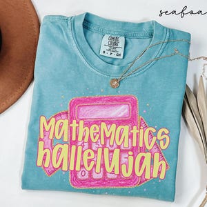 May include: A seafoam green short-sleeve t-shirt featuring a graphic of a pink calculator and a pink protractor. The text "Mathematics hallelujah" is printed in yellow across the center. The shirt has a Comfort Colors brand label at the neck.