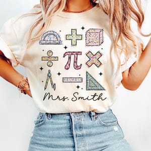 May include: Beige t-shirt featuring colorful math symbols and the name "Mrs. Smith" in a script font. The symbols include a protractor, plus sign, cube, division sign, pi symbol, multiplication sign, compass, line, and triangle.