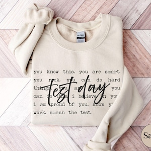 May include: Sand-colored sweatshirt with the words "Test Day" in a handwritten font. The sweatshirt has the phrases "you know this. you are smart" and "smash the test" printed on it. The Gildan brand tag is visible.
