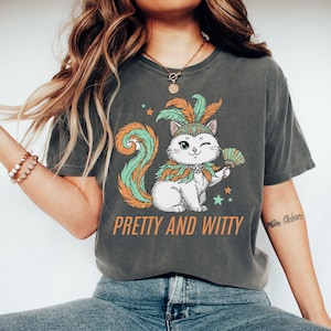 May include: Dark grey t-shirt featuring a cartoon cat wearing a feathered headdress and holding a fan. The cat has a long, swirly tail and is winking. The text "PRETTY AND WITTY" is printed below the cat in orange letters.