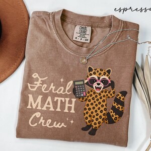 May include: An espresso-colored crew neck t-shirt featuring a graphic of a raccoon with leopard print fur, pink glasses, and a calculator. The text "Feral MATH Crew" is printed in a light cream color next to the raccoon illustration.