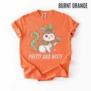 May include: Burnt orange t-shirt with a graphic of a winking cat wearing a feathered headdress and holding a fan. The cat has a green and orange tail and is surrounded by stars. The text "PRETTY AND WITTY" is below the cat.