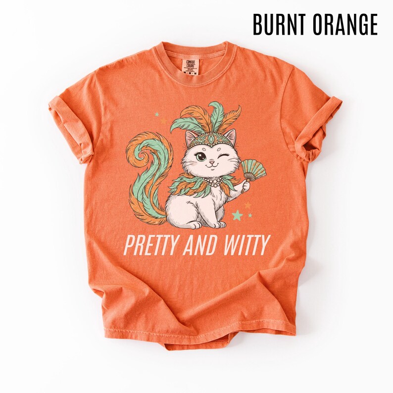 May include: Burnt orange t-shirt with a graphic of a winking cat wearing a feathered headdress and holding a fan. The cat has a green and orange tail and is surrounded by stars. The text "PRETTY AND WITTY" is below the cat.