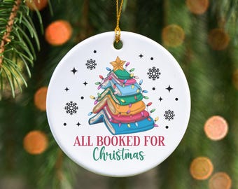 May include: White ceramic Christmas ornament with a colorful stack of books shaped like a Christmas tree. The ornament has the text "ALL BOOKED FOR Christmas" and is decorated with snowflakes and stars.