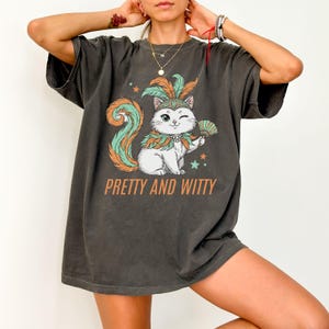 May include: Dark grey t-shirt featuring a cartoon cat with a feathered headdress and fan. The cat has a teal and orange tail and is winking. The text "PRETTY AND WITTY" is printed below the cat in orange.