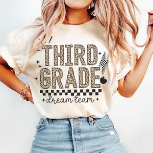 May include: Cream-colored t-shirt with the words "THIRD GRADE" in leopard print. The shirt also features a checkered flag, an apple, and the words "dream team" in a handwritten font.