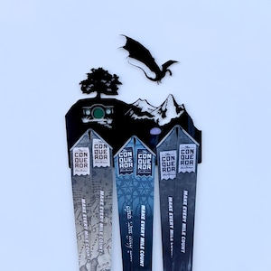 Puede incluir: Tres medallas con temática de El Señor de los Anillos con cintas blancas y negras. Las medallas son plateadas y presentan imágenes de la Comarca, Moria y Rivendel. Las cintas tienen el texto "The Conqueror" y "Make Every Mile Count".